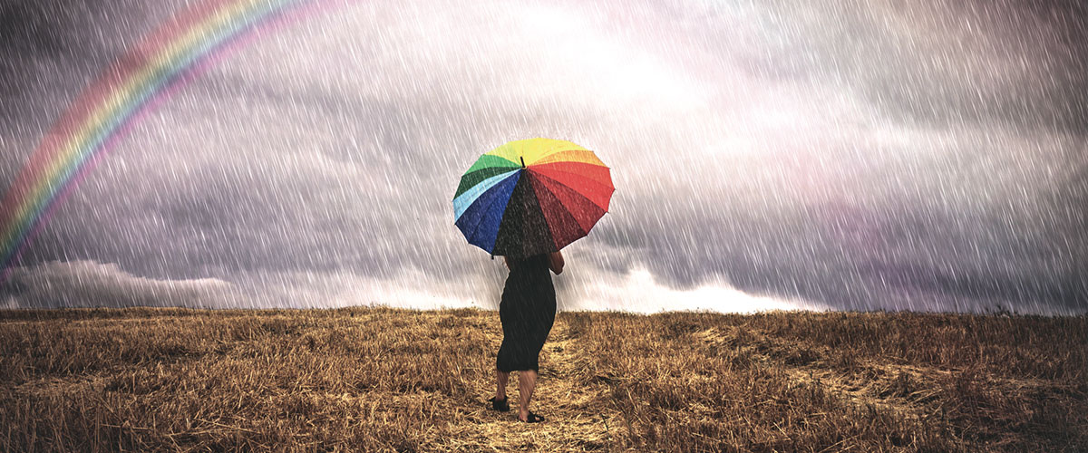 A woman with an umbrella walks under the rain in a field, in the background there is a rainbow. photo WMO