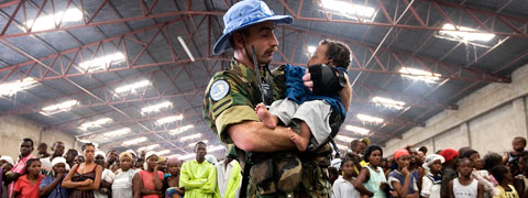 A peacekeeper holding a small child in his arms, standing in front of a crowd of people.