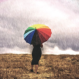 Una mujer con un paraguas camina bajo la lluvia en un campo, al fondo se ve un arco iris. Foto OMM