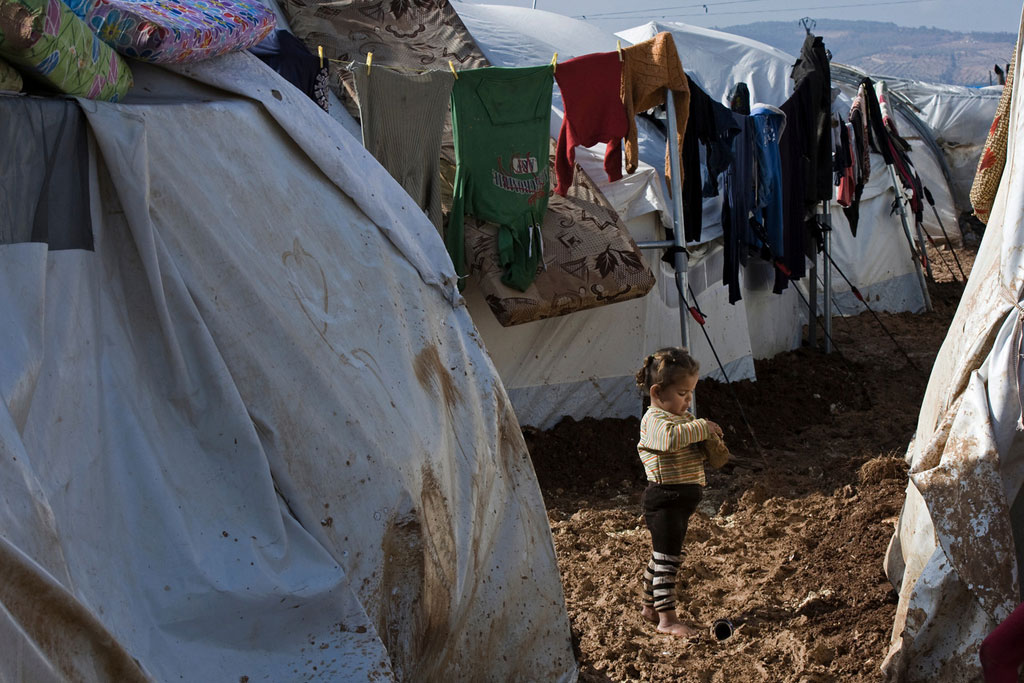 A girl stands barefoot on a muddy walkway between rows of tent shelters, in the Bab Al Salame camp for internally displaced persons, near the border with Turkey, in Aleppo Governorate, Syria. Photo: UNICEF