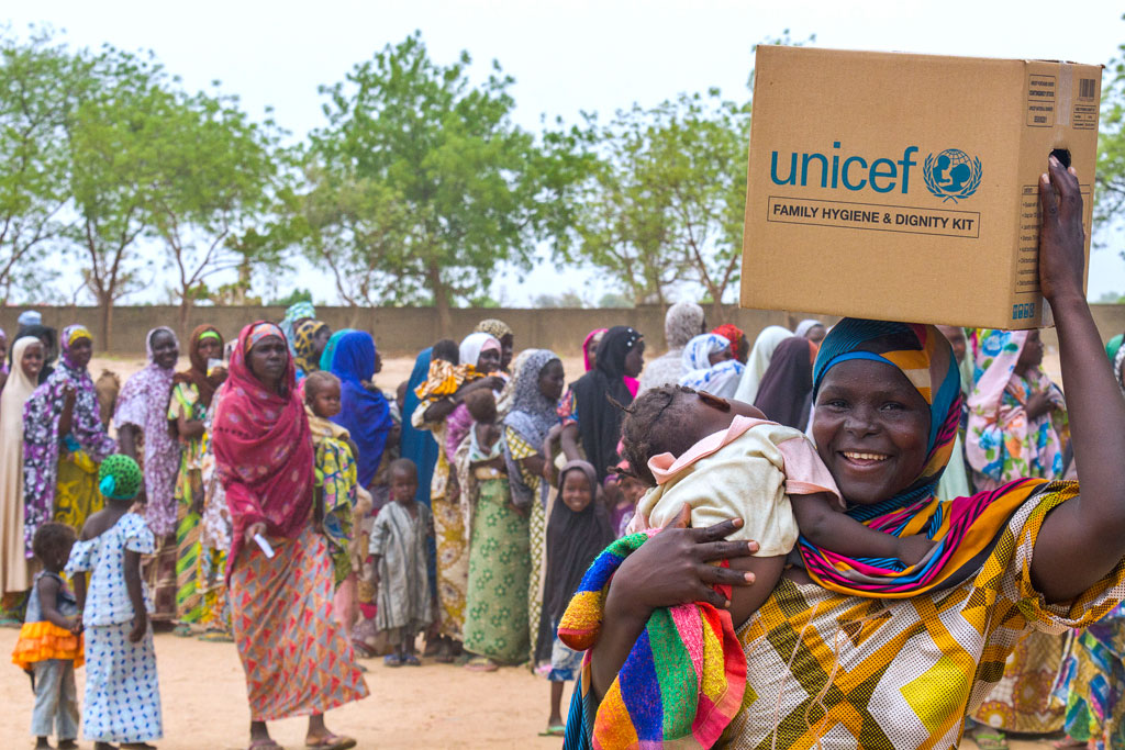 A woman carrying a baby smiles as she leaves a distribution site with a family hygiene and dignity kit, in the Dalori camp for internally displaced people, in the north-eastern Nigerian city of Maiduguri in Borno State. Photo: UNICEF/Andrew Esiebo