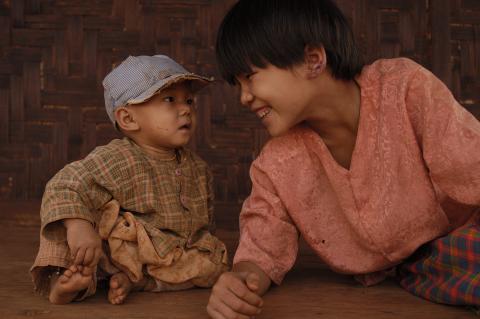 A young girls leans on the ground, smiling at her baby brother, who's seated and wearing a cap.