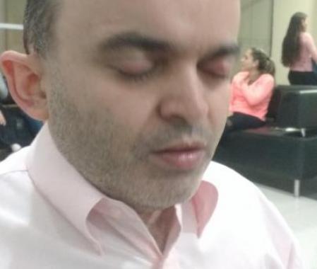 in this picture we can see Alex Garcia, a deafblind Brazilian.  He is with his eyes closed. He has sunglasses on his head, he is wearing a light pink shirt. At the back we can see some people sitting on a sofa. It looks like a hotel/ convention hall