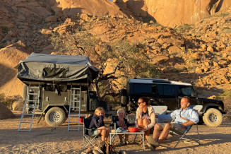 Picture of a family of 4 sat around a camping table in the South African countryside, with a 4x4 and a trailer behind them