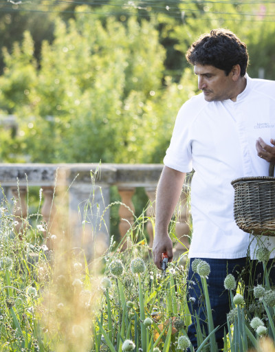 Mauro Colagreco in the garden of the Mirazur, his restaurant in Menton. He is holding a basket filled with fresh herbs on his left arm, and scissors in his right hand as he selects plants to be cooked that day.