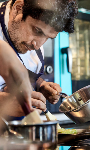 Mauro Colagreco in the kitchen. The Chef very focused on the final touches of a plate; he is leaning over a plate with a brush in his right hand, and holding a saucepan in his left hand. 