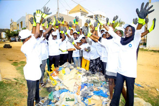 Picture of a group of children in Senegal wearing green gloves and blue caps, standing around a pile of collected waste, smiling and waving their hands in the air
