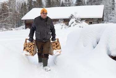 Picture of Tero Mustonen walking through the snow carrying logs in front of a wooden cabin and snowy forest in Finland