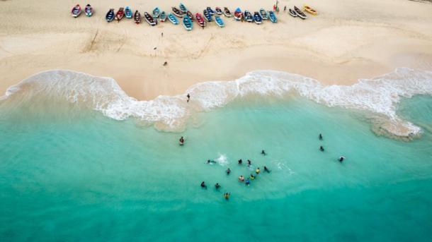 Beach shoreline in Maio Biosphere Reserve, Cabo Verde, with people swimming on a turquoise sea and boats lined up by the sand.